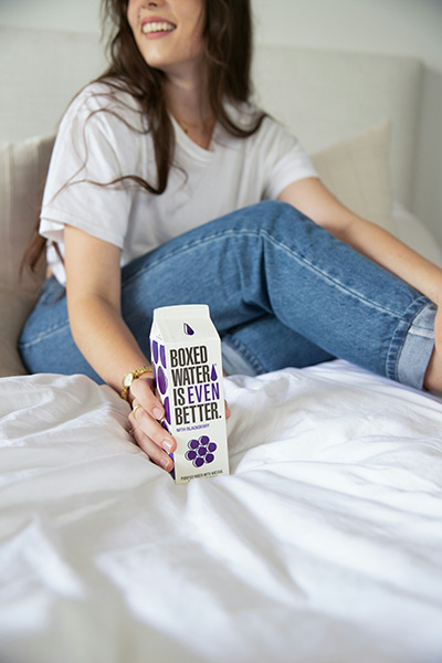 A relaxed woman sits on a bed, holding a carton of boxed water, symbolizing the importance of hydration after a rejuvenating massage session.