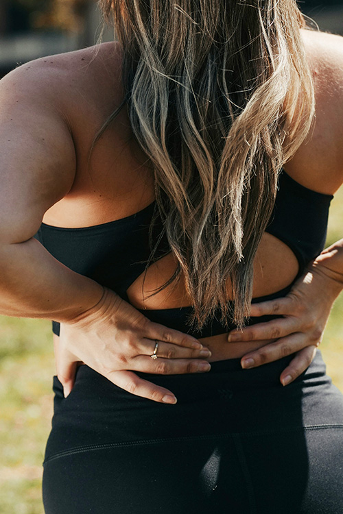 A woman in workout attire appears to be soothing her lower back with her hands, a common gesture for alleviating back pain